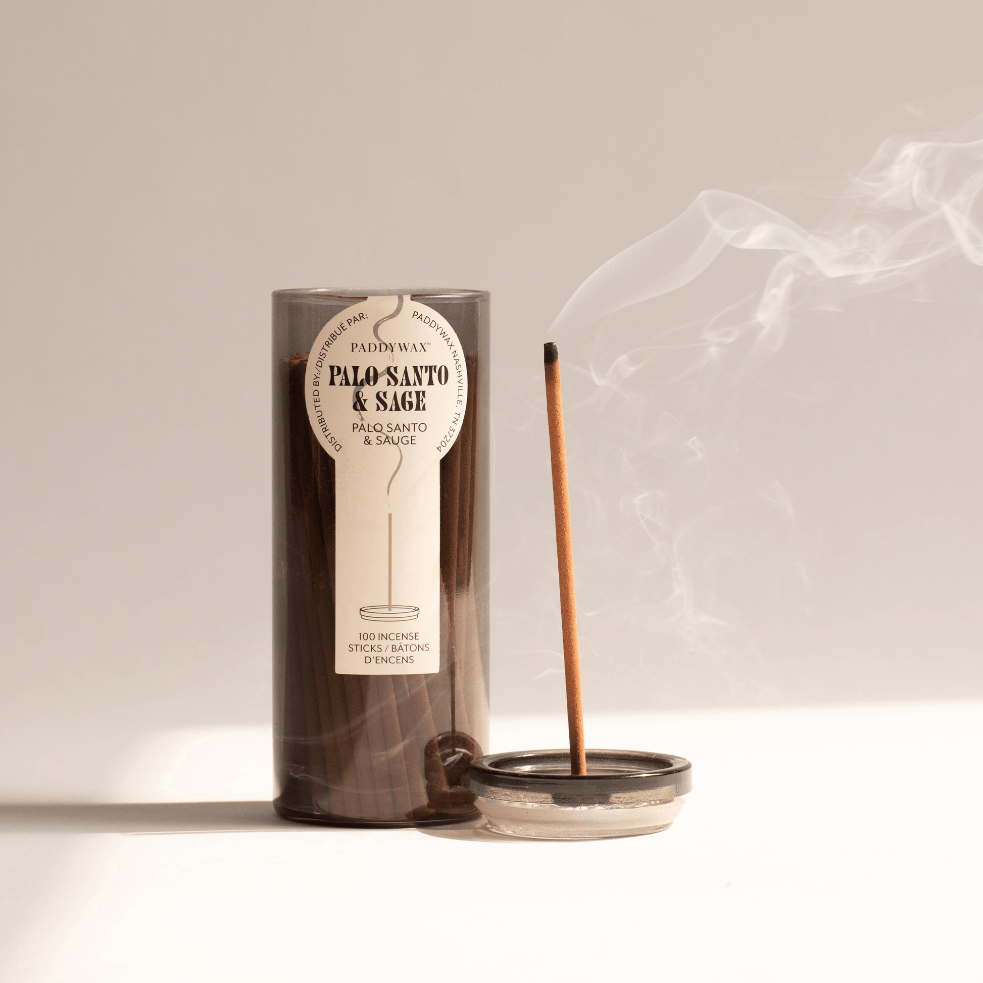 Gray glass cylinder with white label holding 100 incense sticks for flameless fragrance; pictured next to burning incense stick