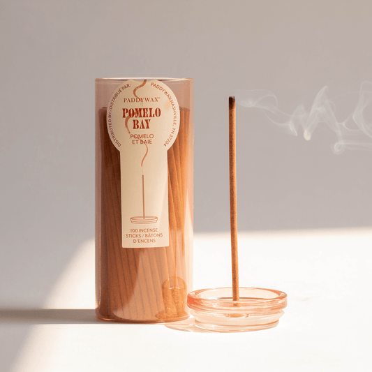 Pink glass cylinder with white label holding 100 incense sticks for flameless fragrance; pictured next to burning incense stick