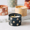The Black Fig + Olive - St. Jude Giveback "Celebrate" 11 oz. Candle sits lit among vibrant ceramic holders—black with smiley faces, yellow with rainbows, sage green with flowers, orange with hearts—all on white surfaces.