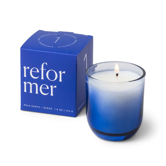 Candle with a vessel of clear glass that fades to blue at the bottom; pictured next to the blue "reformer" box