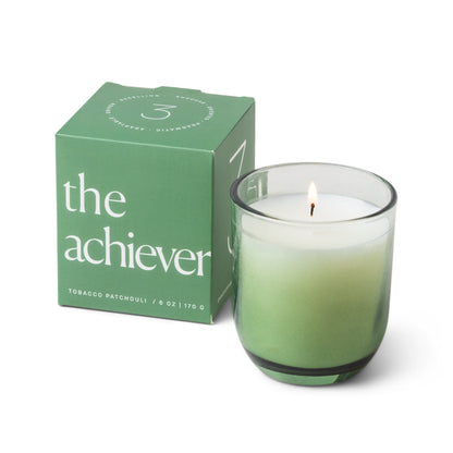 Candle with a vessel of clear glass that fades to green at the bottom; pictured next to the green box which reads “the achiever”