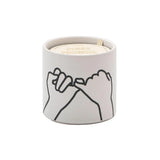The Wild Fig + Cedar "Pinky Promise" Impressions 5.75 oz Candle features a white matte ceramic vessel with a minimalist black line drawing of two hands making a pinky promise and an inner lid that reads PINKY PROMISE.